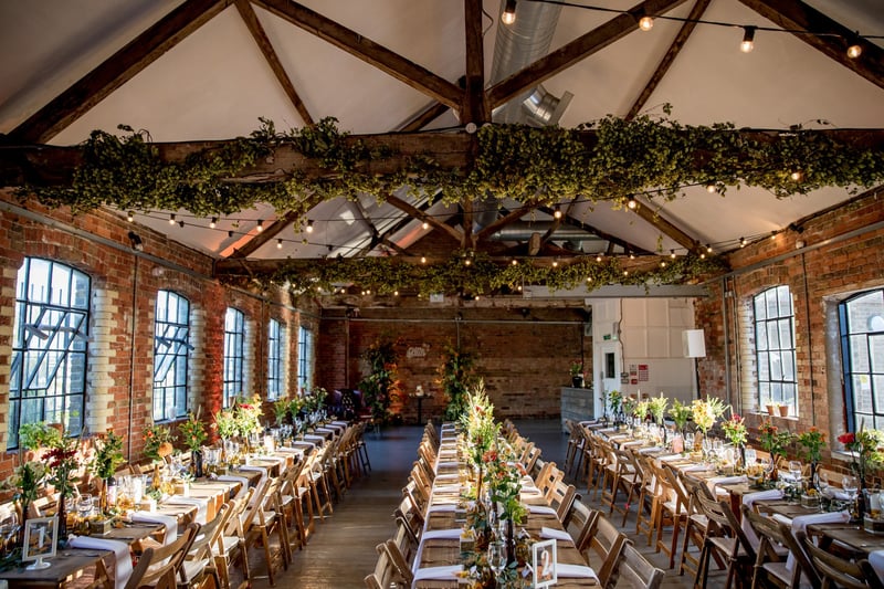 interior of a dry hire wedding venue in london set up for a wedding breakfast with beamed ceiling and long tables filled with foliage.