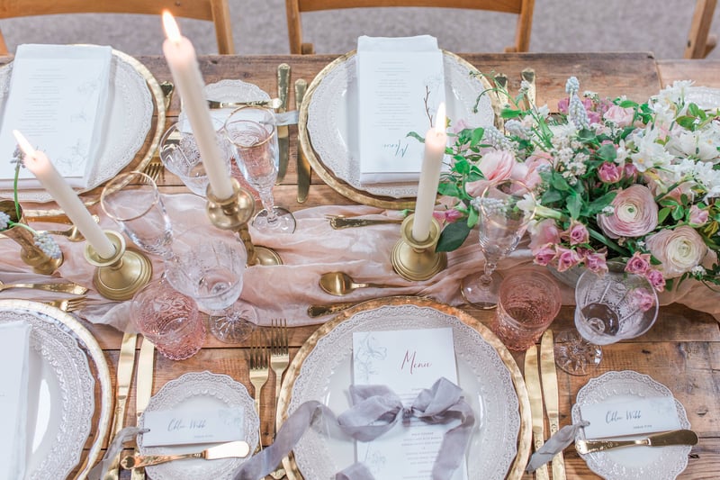 close up of a wedding breakfast table featuring gold-rimmed plates, pink and lilac flowers and a lilac table runner set with taper candles.