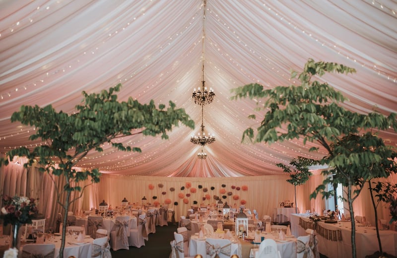 interior of a marquee lined barn with chandeliers and trees, all set up for a wedding breakfast.