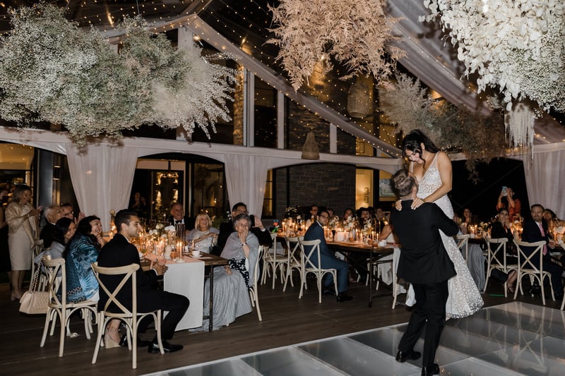 a groom holds his bride in the air during their wedding first dance as seated guests look on from the tables.