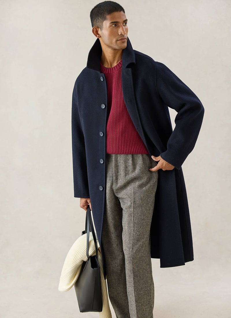 a male model holding a bag wears a navy wool overcoat over grey trousers and a red jumper.