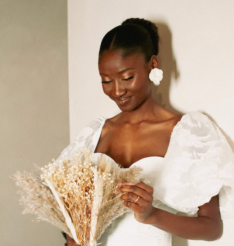 a black bride with a soft glam makeup look and dewy skin looks down at her dried bouquet as she stands in a wedding dress.