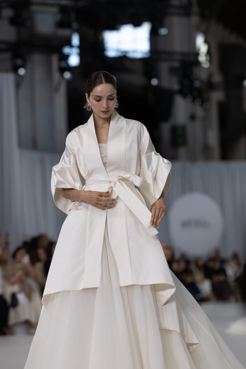a model on a bridal runway wears a silk trench coat with wide sleeves, shawl collar, and waist belt. 