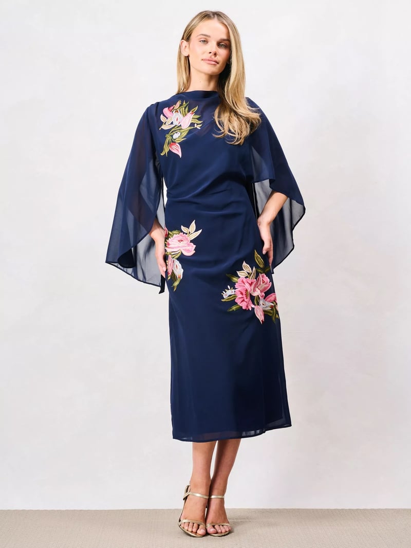 a model wears a navy blue caped midi dress with embroidered pink floral detail.