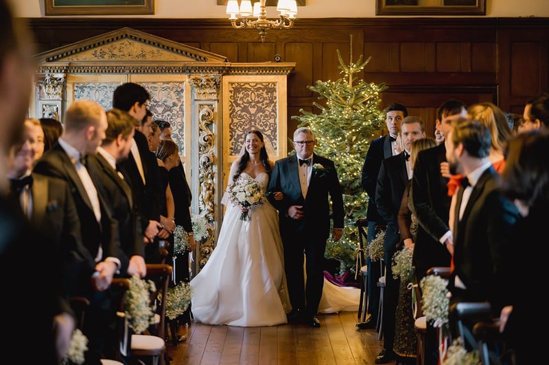 a bride and her father walk down the aisle of a country house wedding venue with a Christmas tree in the background