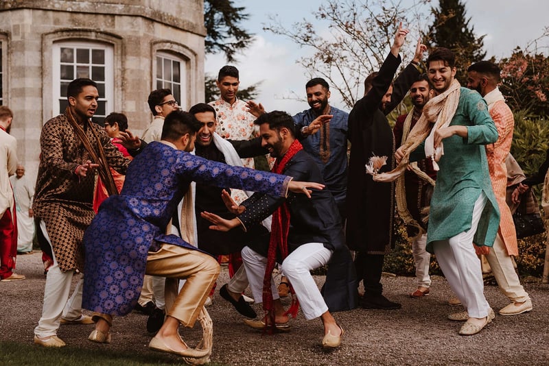 a group of male guests in traditional Hindu wedding attire dance outside a wedding venue.