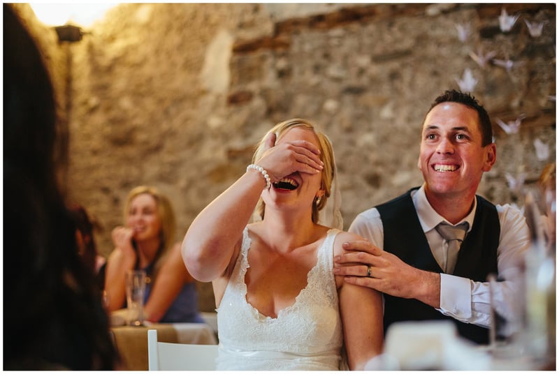 a bride covers her eyes during the wedding speeches as the groom holds her arm and smiles.