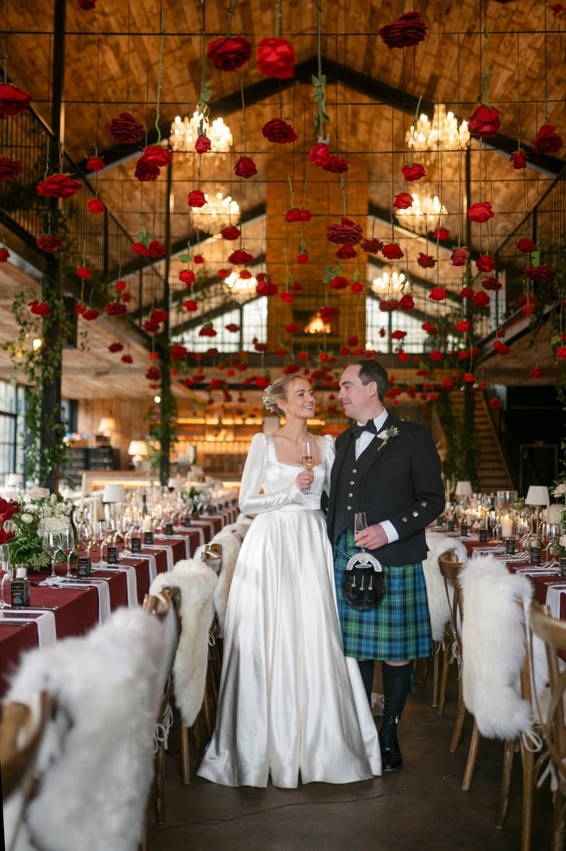 a bride and groom, who is wearing a kilt, smile at each other as they pose in their barn wedding venue with wintery decor.