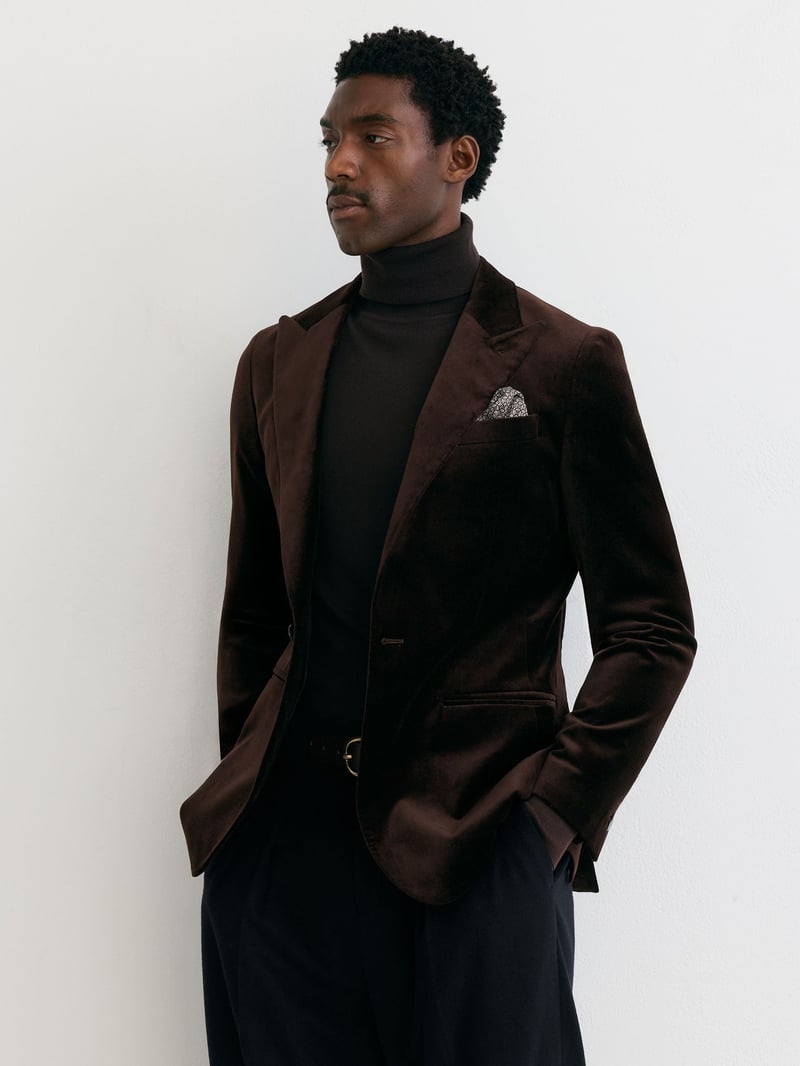 a male model wears a brown velvet blazer with black trousers and a black roll-neck sweater.