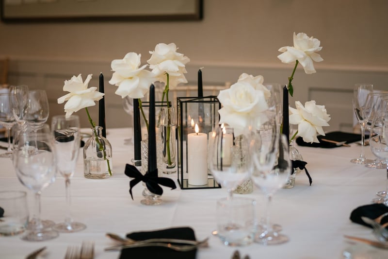 close up of a wedding table with glass vases with single white flowers, glassware tied with black ribbon and pillar candles in hurricane lamps.