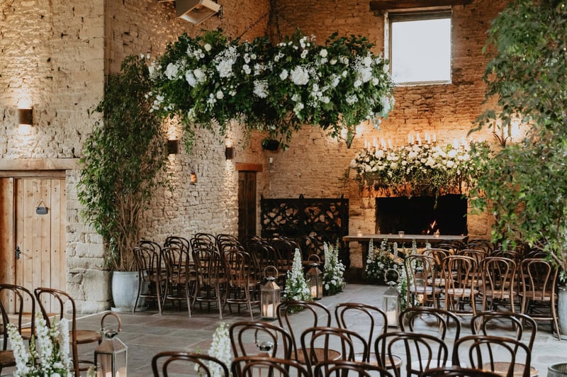 interior of a stone barn wedding venue set up for a ceremony with a floral installation hanging from the ceiling.