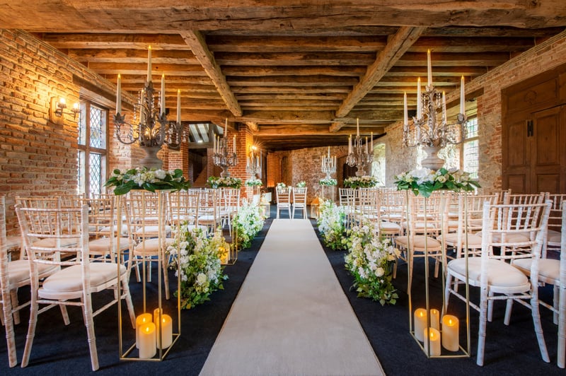 a barn venue set up for a wedding ceremony with candles lining the aisle and white chiavari chairs.