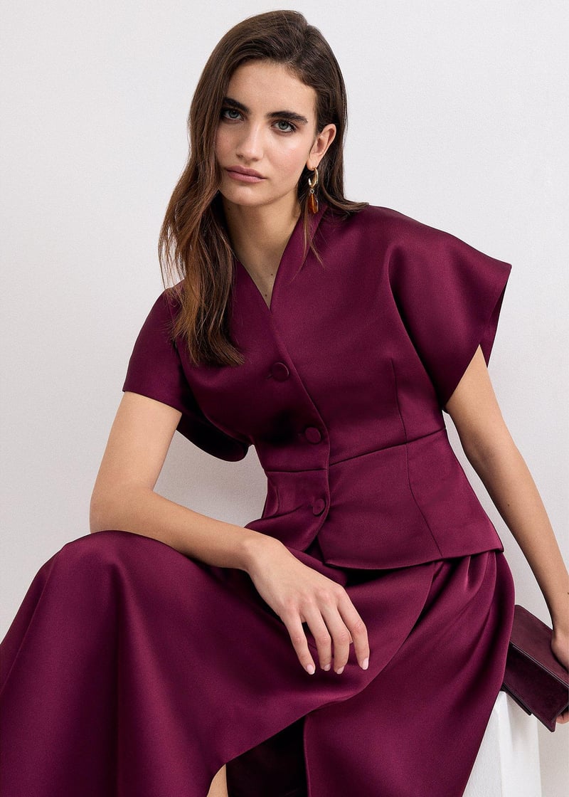 a model sitting down wears a burgundy short sleeved tailored jacket with matching skirt.