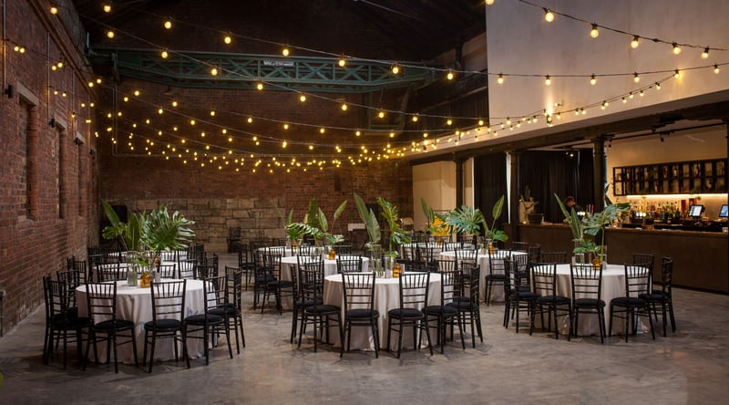 interior of an industrial wedding venue set up for a wedding breakfast with round tables and festoon lighting.