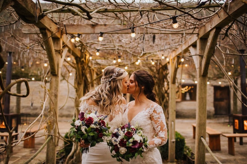 two brides wearing lace dresses and holding winter red and white bouquets kiss under a fairy-lit pagoda.