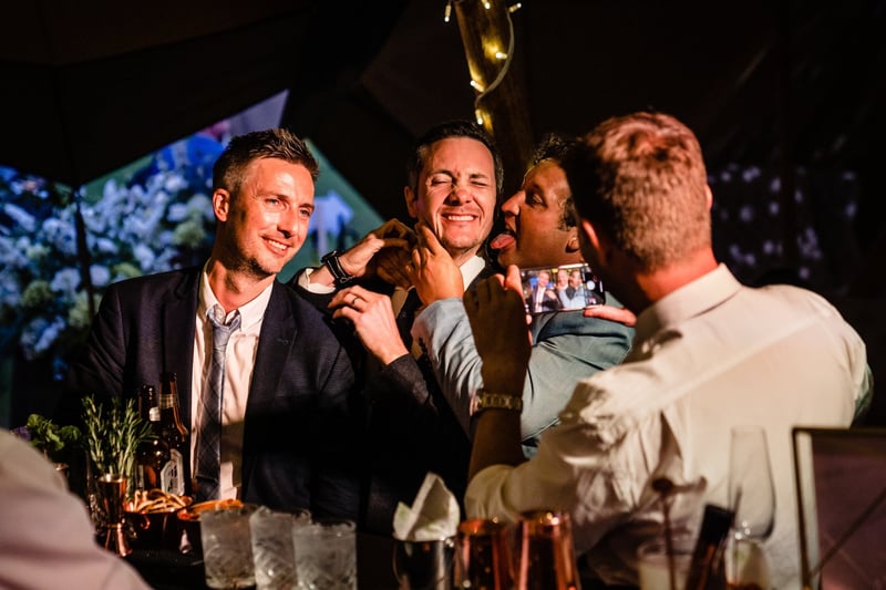 a group of male wedding guests joke around at the bar as one guest takes a photo on his phone.