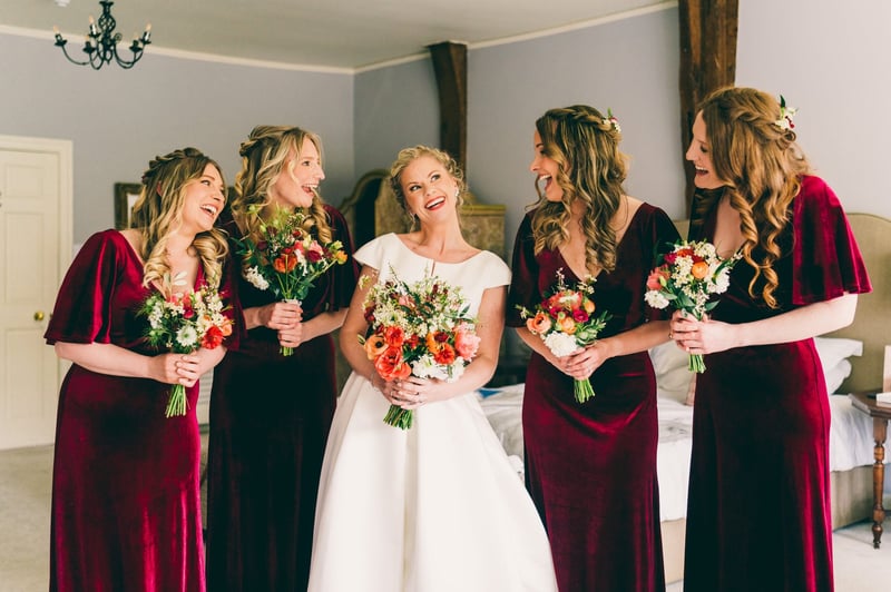 a bride and her bridesmaids, who are wearing red velvet dresses, smile as they pose with their Christmas wedding bouquets.