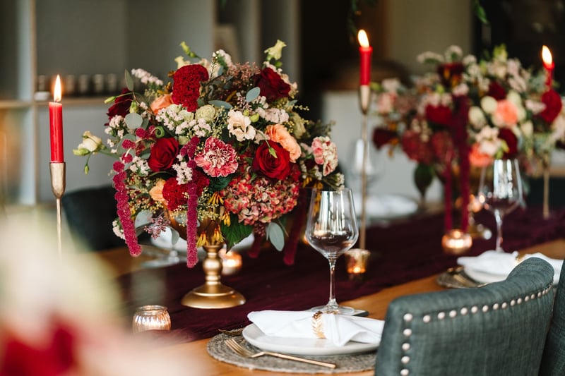 close up of a gold urn centrepiece on a table with tall candles, featuring red, cream and green flowers and foliage, including trailing amaranthus.