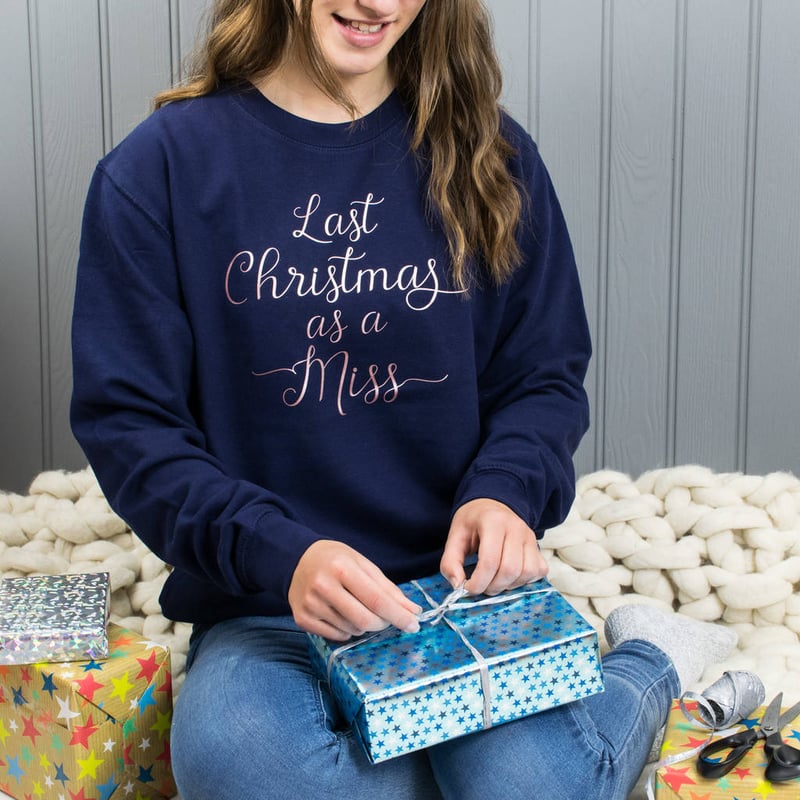 a model opening a gift wears a navy blue Last Christmas as a Miss sweatshirt with rose gold script writing.