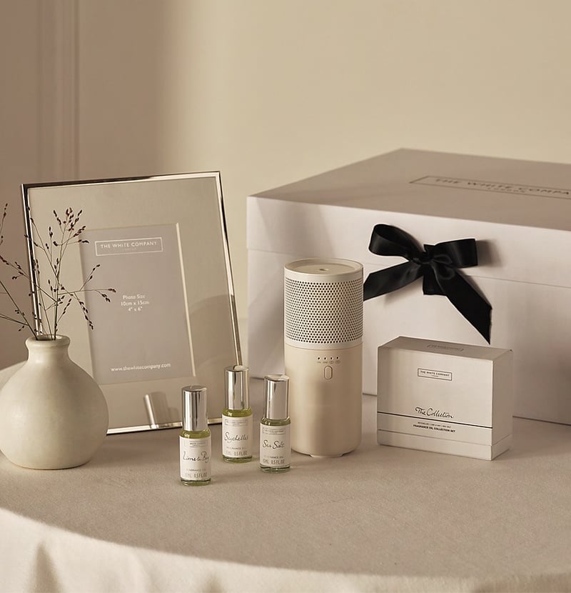 an arrangement of gift items on a table, including a diffuser and photo frame next to a White Company signature box tied with black ribbon.
