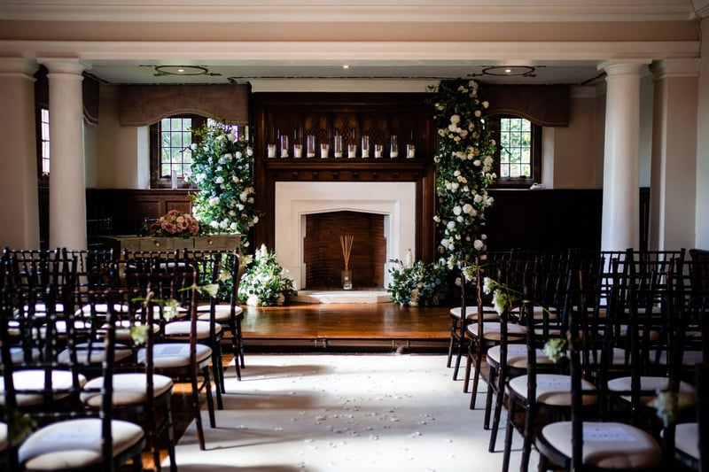 interior of a country house wedding venue set up for a ceremony in front of a fireplace with wood panelling and foliage.