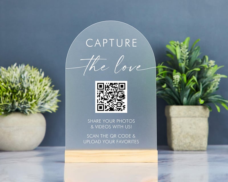 an acrylic QR code sign for wedding on a table with potted plants against a grey wall. 