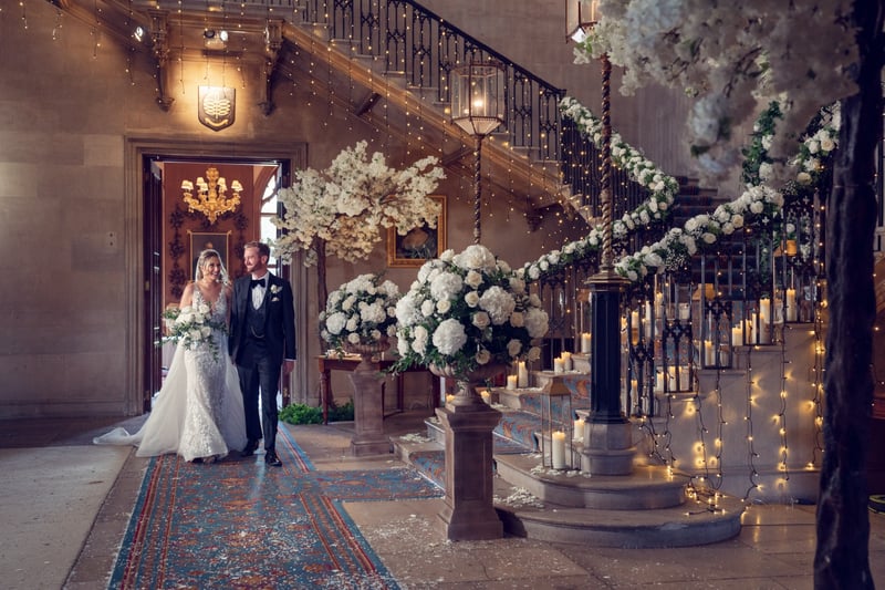 a bride and groom walk through the marble hall of a country house wedding venue decorated with white flowers and candles.