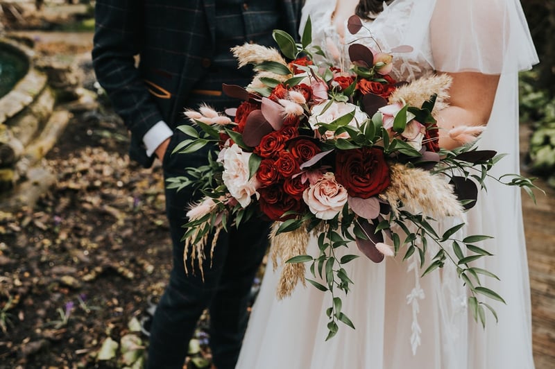 close up of a bride holding a wild and natural winter wedding bouquet with red roses, greenery and blush coloured ranunculus.