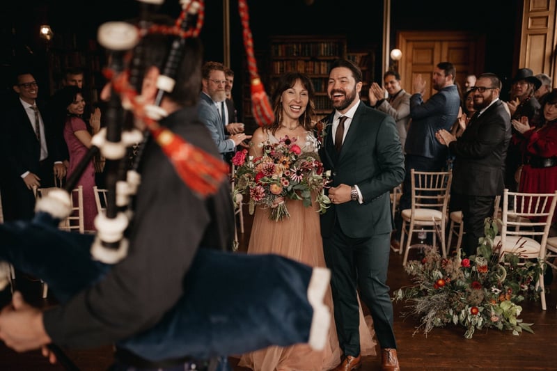 a bride and groom in a cosy wood-panelled room smile as they watch a bagpiper perform at their winter wedding ceremony. The bride is holding a wintery bouquet and there are floral arrangements at each seat end.