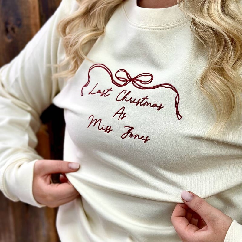 close up of a woman wearing a cream sweatshirt with red bow embroidery and writing.
