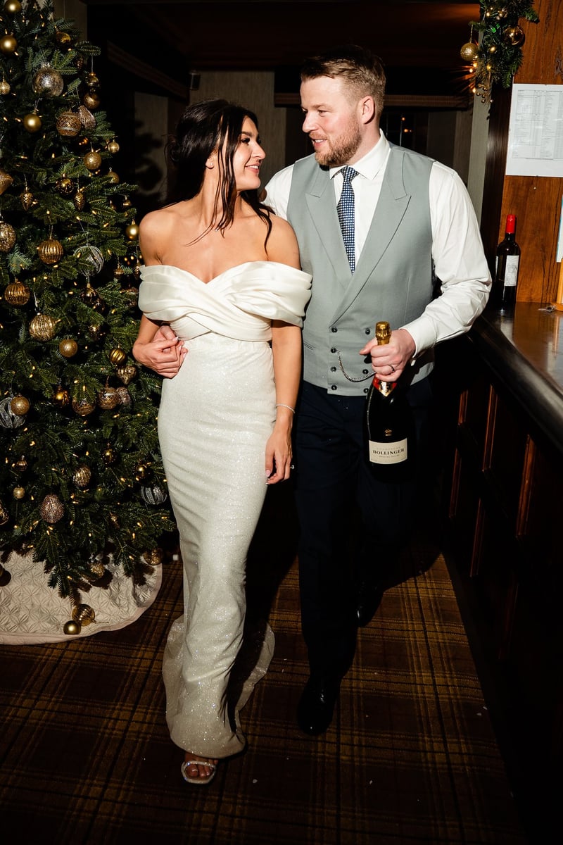 a bride and groom kiss as they walk through their wedding venue clutching a bottle of champagne. There is a Christmas tree in the background.