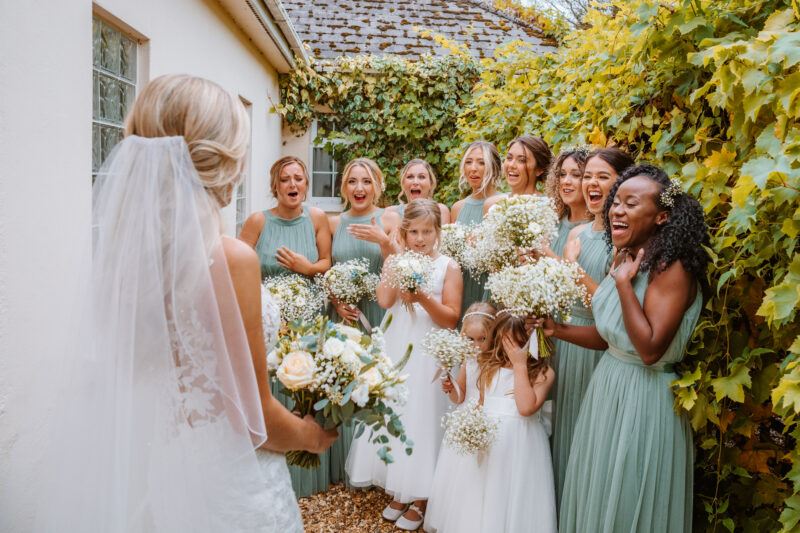 A bride stands on the left with her back to the camera. A group of bridesmaids in green and white dresses and holding bridal bouquets react to her entrance. 