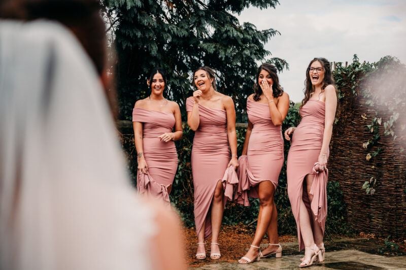 The back of a bride's head appears on the left out of focus with her white veil visible. A group of four bridesmaids in pink dresses stand in a row and react to the bride's entrance with laughter and smiles.