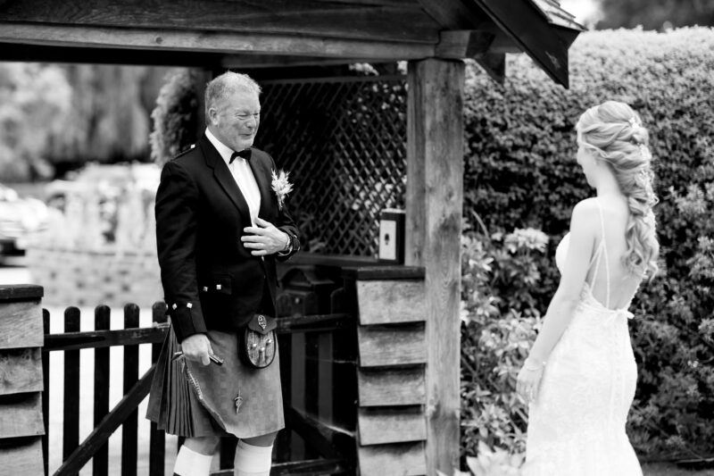 The father of the bride stands on the left by a gate as his daughter the bride comes towards him in her wedding dress with her back to the camera. He places his hand on his chest as he proudly admires her.