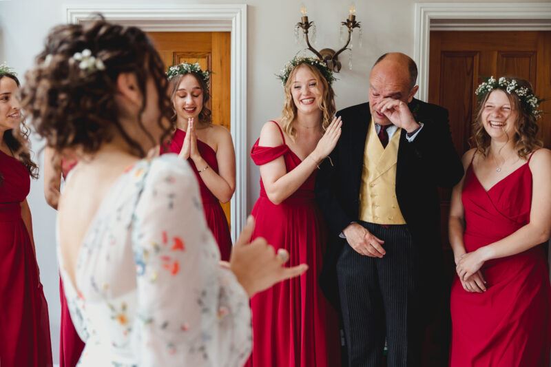 A bride stands with her back to the camera in a floral wedding dress. Her father stands facing her with the bridesmaids, each wearing red dresses and smiling, wiping a tear from his eyes.