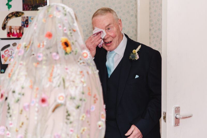 A bride's floral veil appears from behind on the left, as the father of the bride stands in the centre, face to the camera, wiping a tear from his eye as he admires his daughter in her wedding dress for the first time.