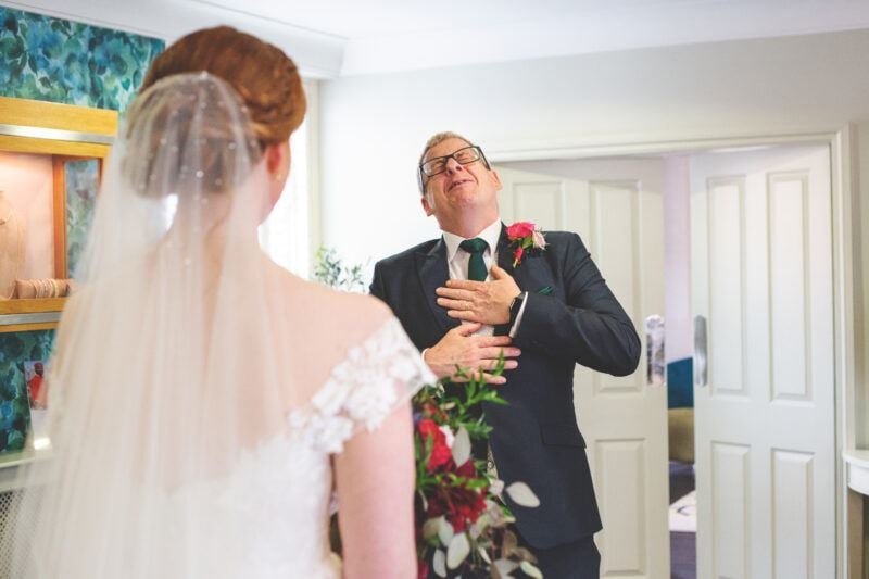 A bride stands on the left in her wedding dress, facing her father who clutches his chest with both hands, completely overcome with emotion at the first look.