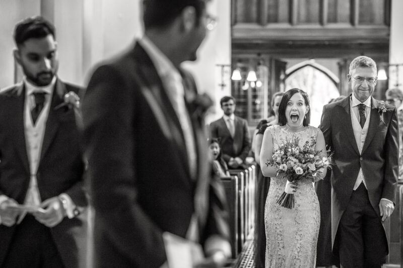 A groom stands in the centre of the photo but out of focus, turning round to face his bride as she walks down the aisle, her mouth wide open in shock and excitement, accompanied by her father, surrounded by guests in the church.