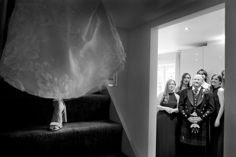 On the left a bride steps down the stairs, just her high-heel shoe and the bottom of her wedding dress visible, while on the right, her father and bridesmaids stand awaiting her entrance in anticipation for the first look.