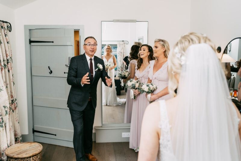 A bride stands with her back to the camera in a dressing room, as her father enters through a sage-green door for the first look, his hands spread wide in surprise, while two bridesmaids stand in the background to the left, both smiling and holding bridal bouquets.