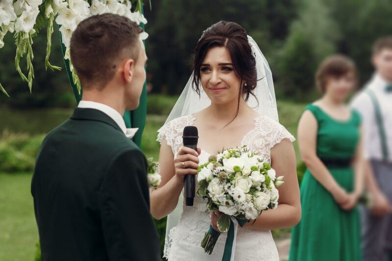 Gorgeous emotional bride performing her vows wearing a stylish white wedding dress with bouquet taking vow during outdoor wedding ceremony near aisle to handsome groom, her eyes tearing up