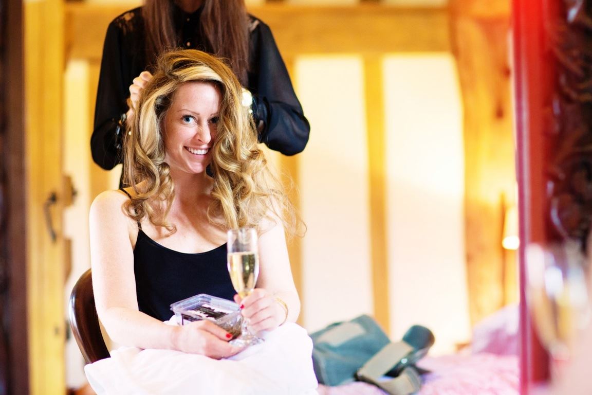 Bridebook.co.uk- bride with a glass of champagne having her hair styled