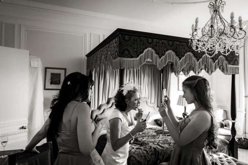 www.bridebook.co.uk bride applying lipstick as bridesmaids hold up a mirror