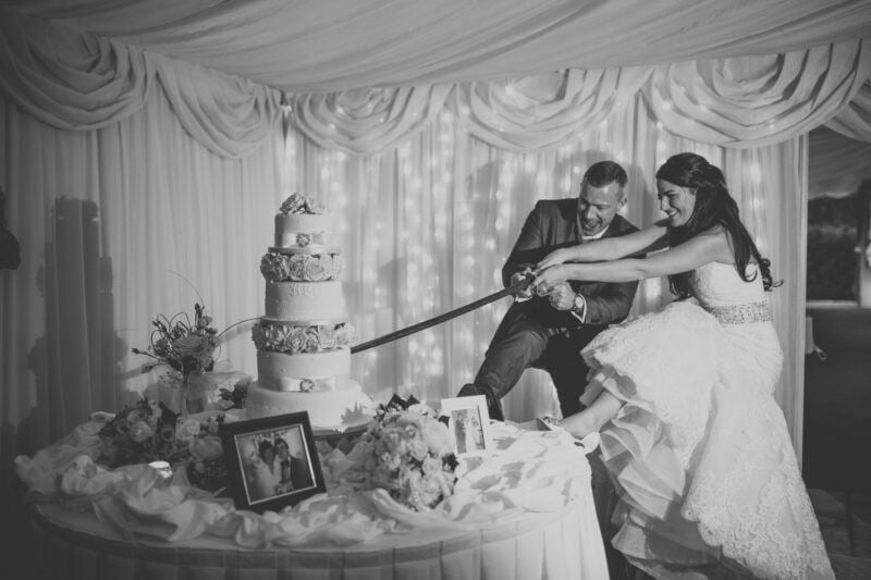 Bridebook.co.uk- bride and groom having fun cutting the cake with a ceremonial sword