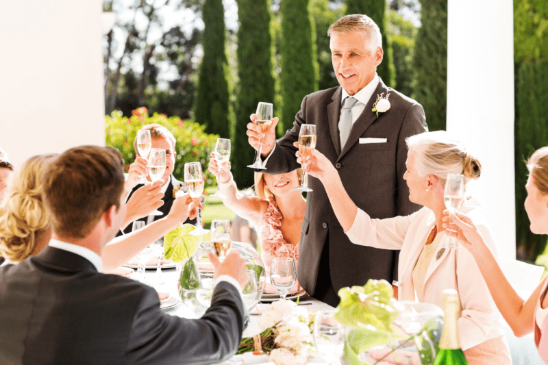 Father of the bride raising a glass to toast the bride and groom with other guests raising their glasses too