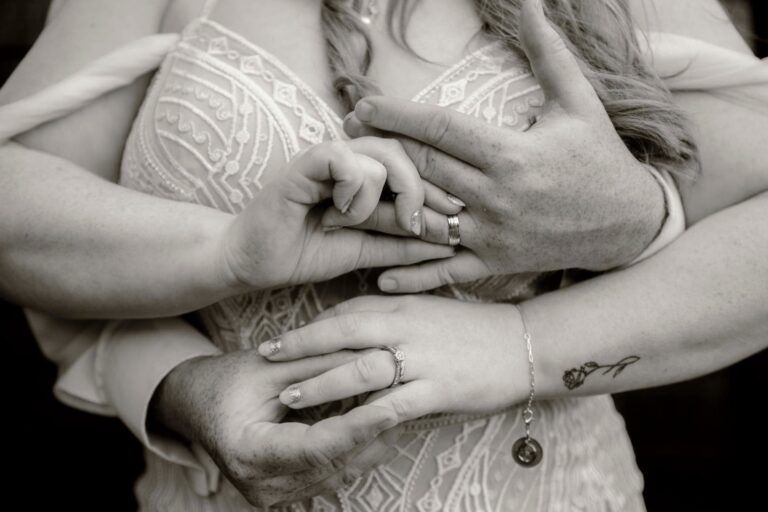 Front view of bride, embraced by groom from behind, their hands clasped in front, showing off engagement ring
