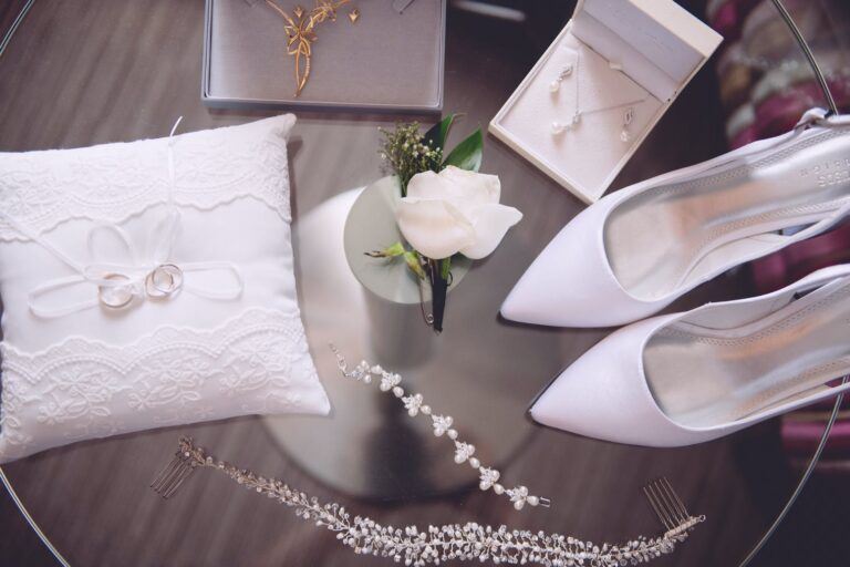 Assortment of wedding accessories, including engagement ring and wedding rings on white pillow, white shoes, and jewellery