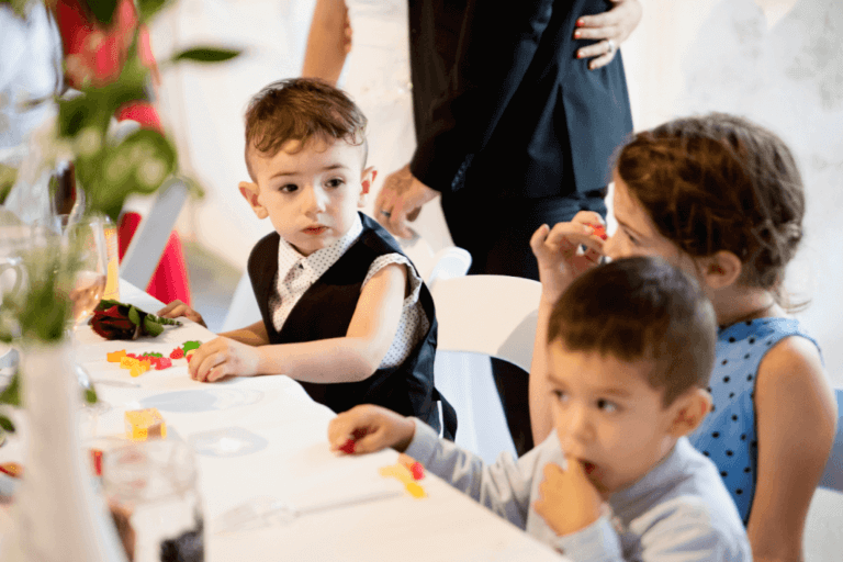 Three young children are seated at a table during a formal wedding event. The children are engaged with colorful toys and a few snacks. The child on the left is wearing a vest and looking to the side, while the other two children focus on their toys.