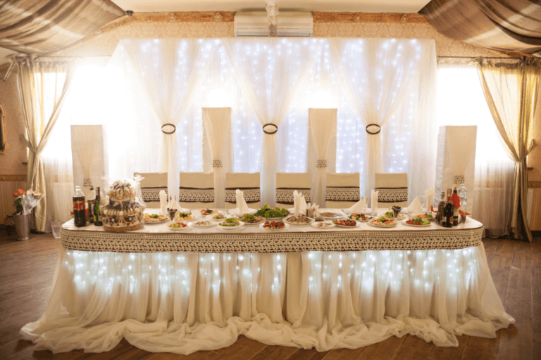 A beautifully decorated wedding table adorned with white drapes and illuminated with small lights. The table is set with plates of food, drinks, and napkin holders, with high, white cushioned chairs behind it. The background features soft, glowing curtains.