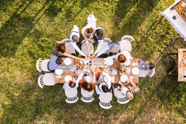 Aerial view of a group of people seated at a long outdoor table, sharing a meal. The table is set with plates, cutlery, various dishes, and flowers. The gathering takes place on a grassy lawn, with sunlight casting shadows around the scene.